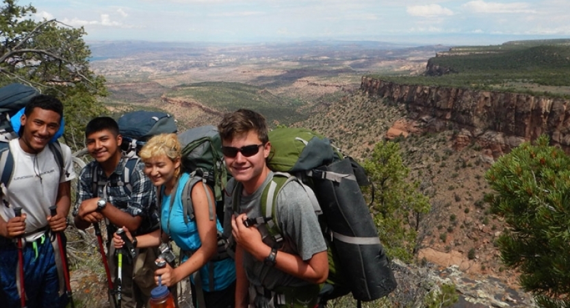 Four students wearing backpacks smile at the camera. They are high above a southwestern landscape.